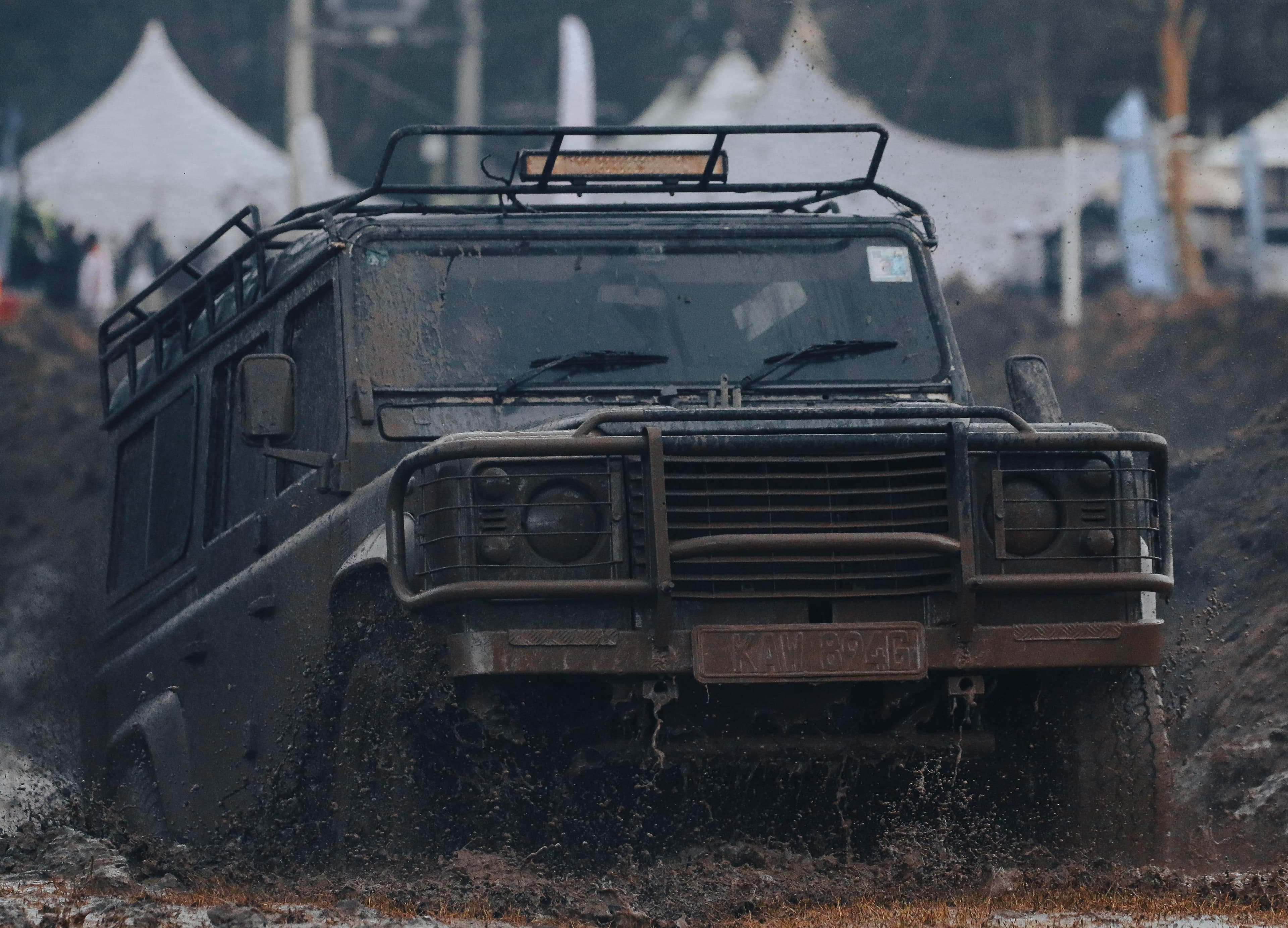 A mud-caked 4x4 crossing the course with a row of white camping tents pitched along the tree line behind it